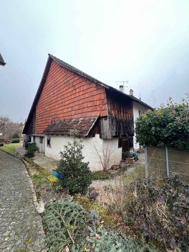 A rustic two-story house with a red-tiled roof and white walls, surrounded by greenery and a stone pathway.