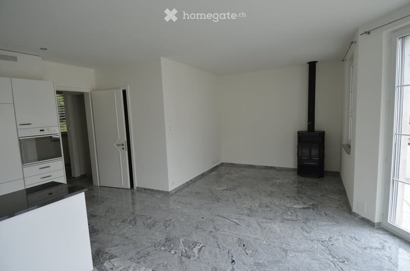Empty room with white walls, marble flooring, and a fireplace in the corner. There is a kitchen area with white cabinets and a countertop visible on the left side.