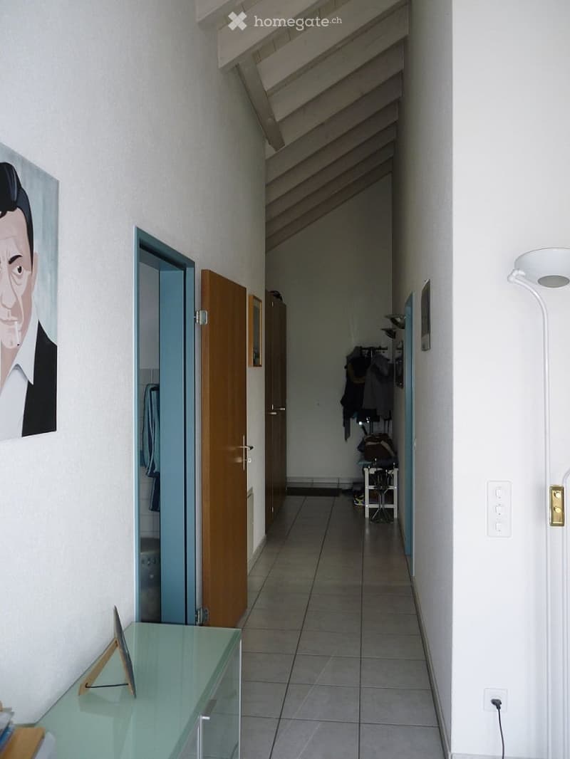 A narrow hallway with tiled flooring, white walls, and a slanted ceiling. Features include a glass console table with a small decorative item, a painting on the wall, and several doors leading to other rooms.