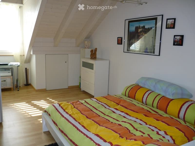 Bright bedroom with wooden floors, slanted ceiling beams, and a colorful striped bedspread. Includes a nightstand, mirror, and decorative items on a cabinet.