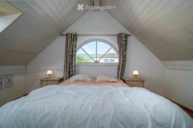 Cozy attic bedroom with slanted wooden ceilings, large arched window, bed with white duvet and colorful pillows, two nightstands with lamps, and patterned curtains.