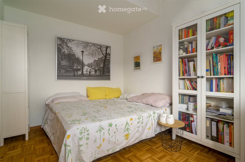 Cozy bedroom with a single bed, colorful pillows, a large bookshelf filled with books and decorative items, a small round stool with two white ceramic figures, and framed artwork on the walls.