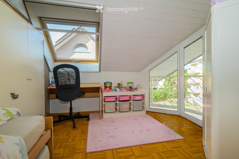 Bright bedroom with wooden flooring, slanted white ceiling, large window, and open balcony door leading to a terrace. Contains a bed, desk with chair, storage unit with bins, and a pink rug.
