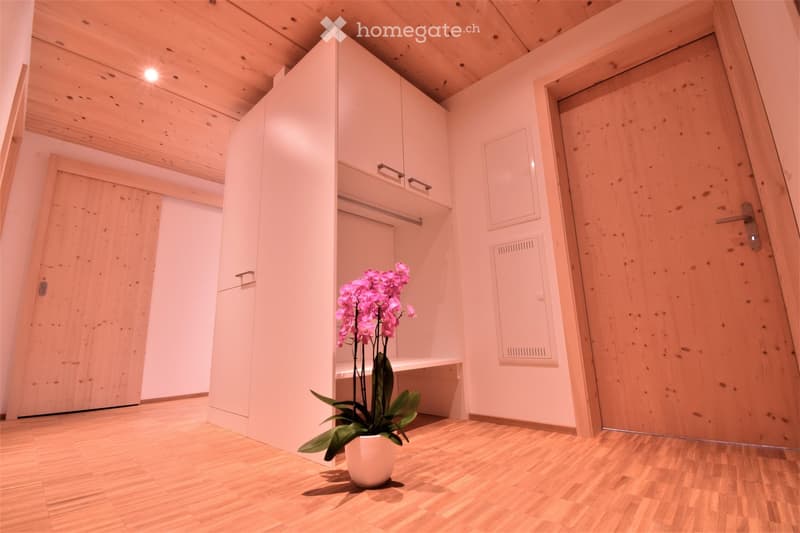 A small, modern hallway with wooden flooring, white walls, and a built-in wardrobe. A potted orchid is placed on the floor near the wardrobe.