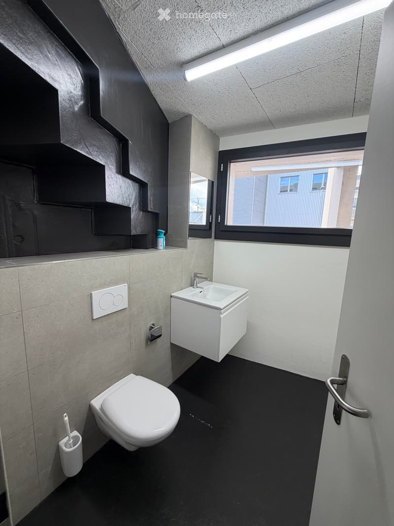 Modern bathroom with a toilet, sink, and mirror. Features dark grey tiles, white fixtures, and a window with a view of the outside.