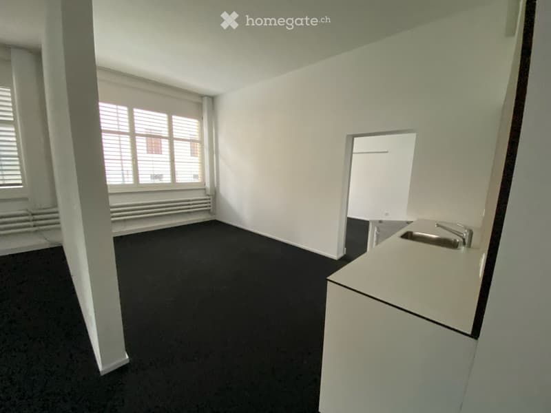 Empty room with white walls, black carpet, and a small sink in the corner. Windows with blinds and a radiator are visible.