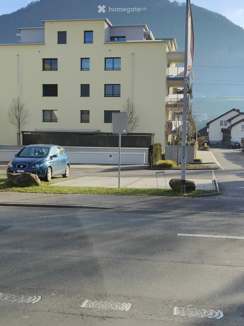 A multi-story residential building with light yellow walls and multiple windows, featuring a covered parking area and a small garden area.