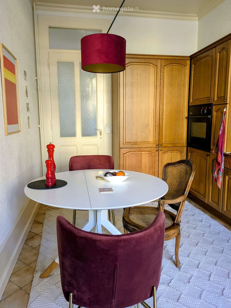 A cozy kitchen with a round white dining table, red chairs, a wicker chair, a bowl of fruit, a pepper grinder, wooden cabinets, and an oven.