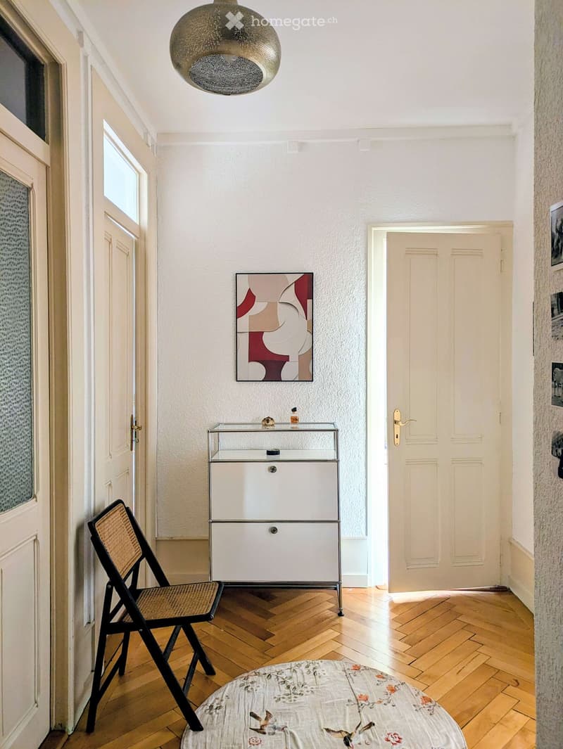 A narrow hallway with wooden flooring, a hanging spherical light fixture, a small white cabinet with glass top, a round rug with floral pattern, and a folding chair.