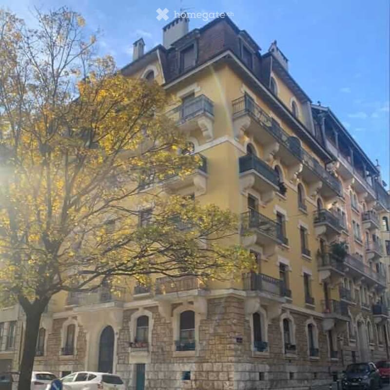 A multi-story residential building with yellow facade, multiple balconies, and ornate architectural details. The building has a stone base and is surrounded by trees and parked cars.
