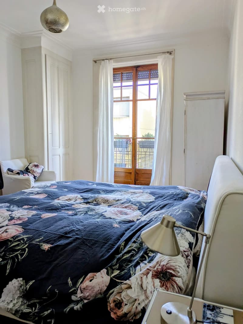 Cozy bedroom with a large bed covered in a floral-patterned duvet, a nightstand with a lamp, a wooden door leading to a balcony, and a small armchair with a patterned pillow.
