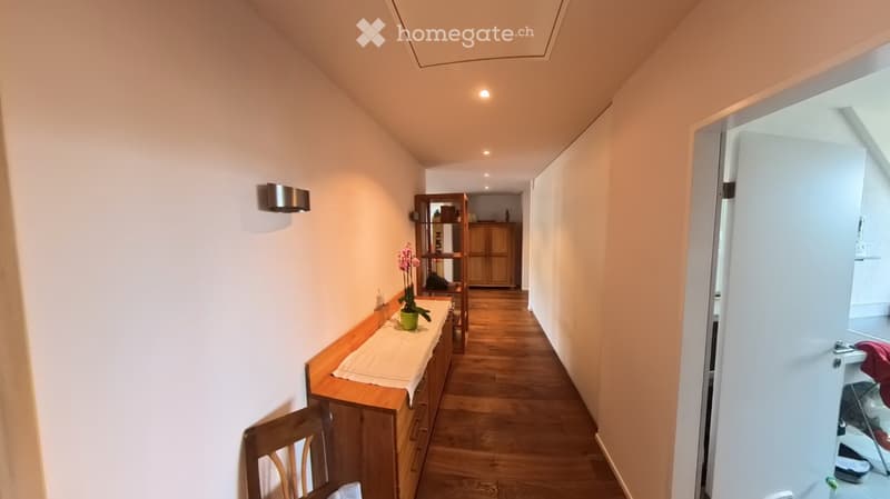 A narrow hallway with wooden flooring, light-colored walls, and built-in storage units. A small table with a vase of flowers is visible on the left side, and a partially open door leads to another room on the right.