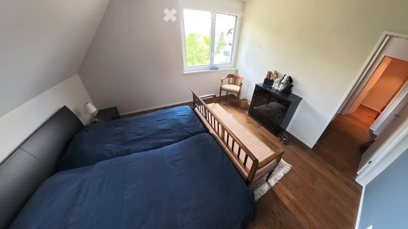 Cozy attic bedroom with a blue sofa, wooden chair, dresser, and table lamp. The room features wooden flooring and large windows with a view of greenery.