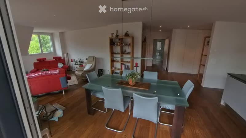 Modern living and dining area with wooden flooring, a red sofa, a glass dining table with chairs, and a bookshelf with decorative items.