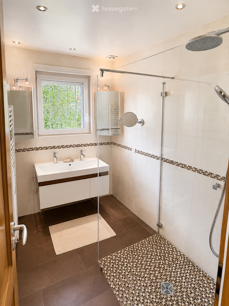 Modern bathroom with a large window, double sink, shower area, and built-in storage. Features include a glass shower enclosure, a large mirror, and a tiled floor.