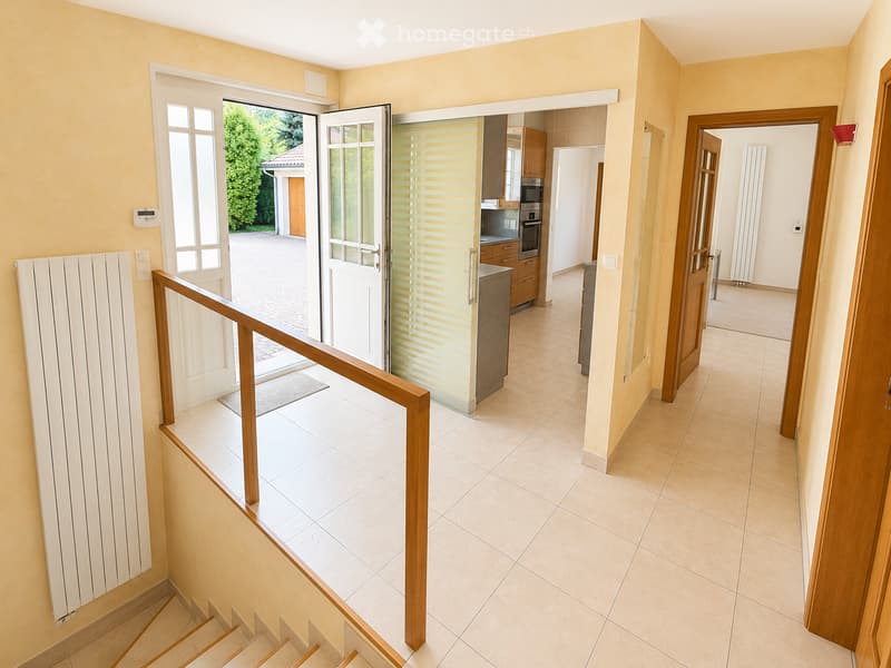 Spacious hallway with tiled flooring, wooden staircase, and open doors leading to a kitchen and outdoor area.