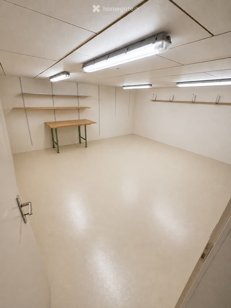 Empty room with beige walls, wooden floor, and built-in wooden shelves. A small wooden table with green legs is placed against one wall. The room has fluorescent lighting fixtures on the ceiling.
