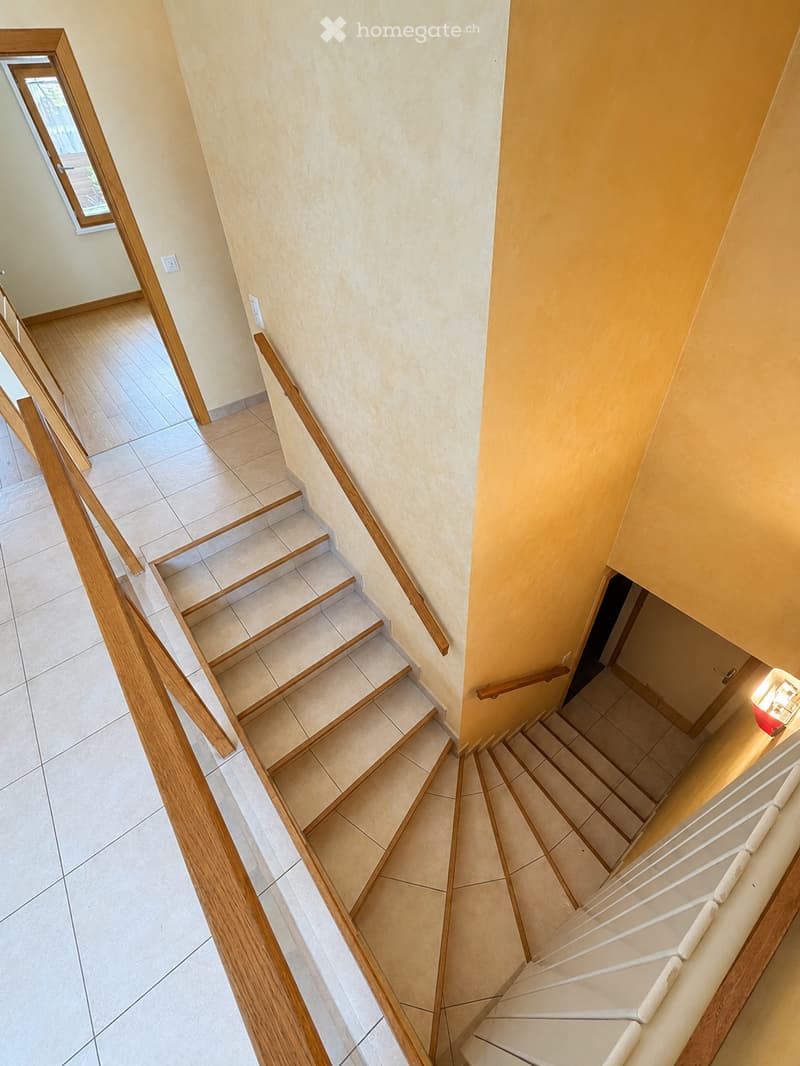 Spacious hallway with wooden staircase featuring beige tiles and wooden railings, light-colored walls, and a small window at the top left.
