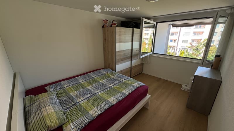 Cozy bedroom with a single bed, wardrobe, nightstand, and mirror. The room features wooden flooring and large windows with a view of the balcony and surrounding buildings.