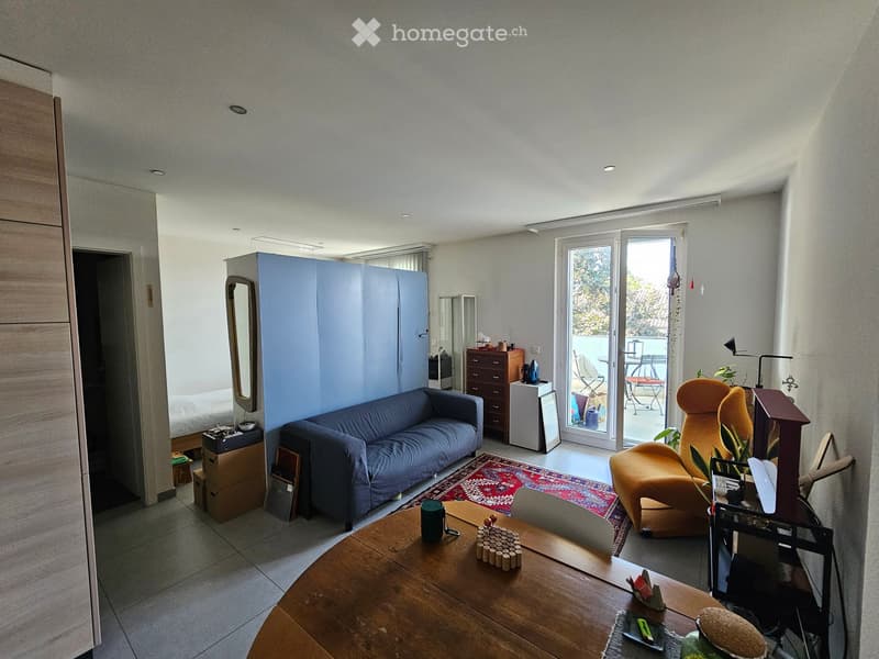 A modern living room with a blue sofa, orange armchair, wooden coffee table, and a view to a balcony. The room features a large sliding glass door leading to the balcony, a patterned rug, and a small cabinet with a mirror.