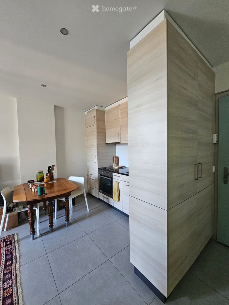 Modern kitchen with light wood cabinets, a wooden dining table with chairs, and various kitchen items on the table.