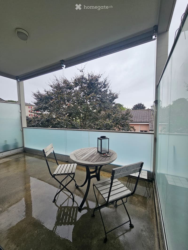 Balcony with a small round wooden table, two folding chairs, and a lantern, overlooking a residential area with trees and houses.