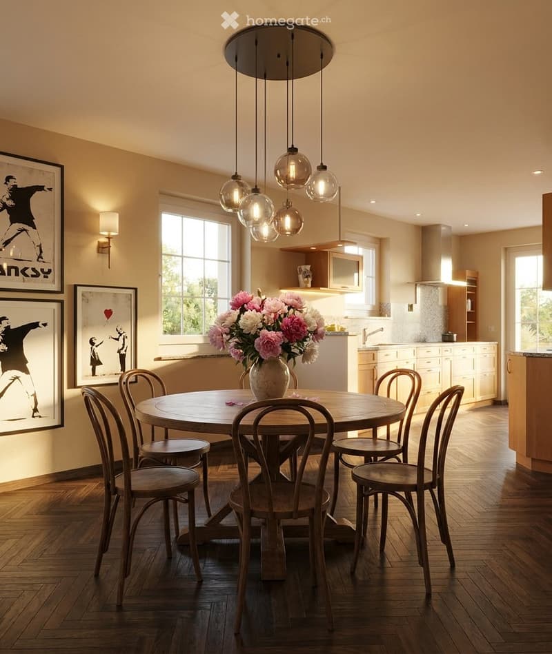 Open-plan dining and kitchen area with a round wooden table surrounded by six wooden chairs, a vase of pink and white flowers, framed artwork on the walls, and a modern pendant light fixture above the table. The kitchen features white cabinets, a sink, an