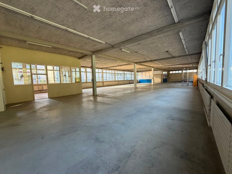 Large, empty industrial-style room with concrete floors, exposed ceiling beams, and large windows allowing natural light. Minimal furnishings, with a few scattered items like a ladder and small cabinets visible in the background.