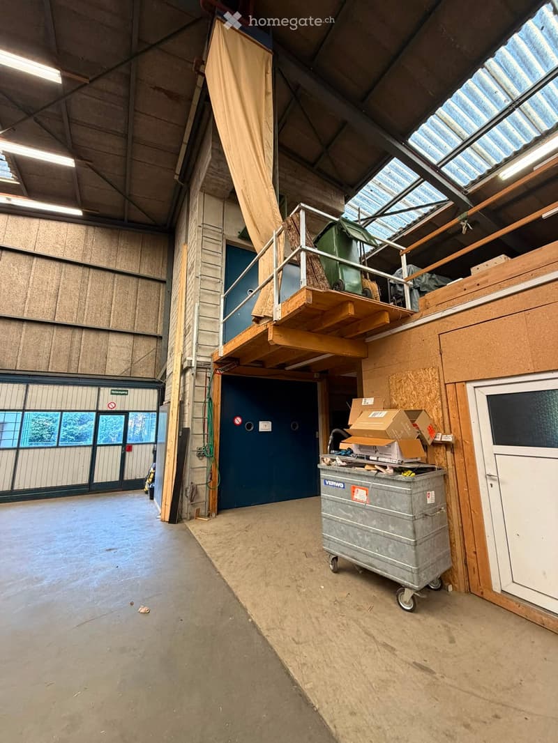 Empty industrial-style room with concrete floor, exposed wooden beams, and large windows. Features a metal staircase leading to an upper level, a large blue door, and a grey wheeled container with boxes on top.