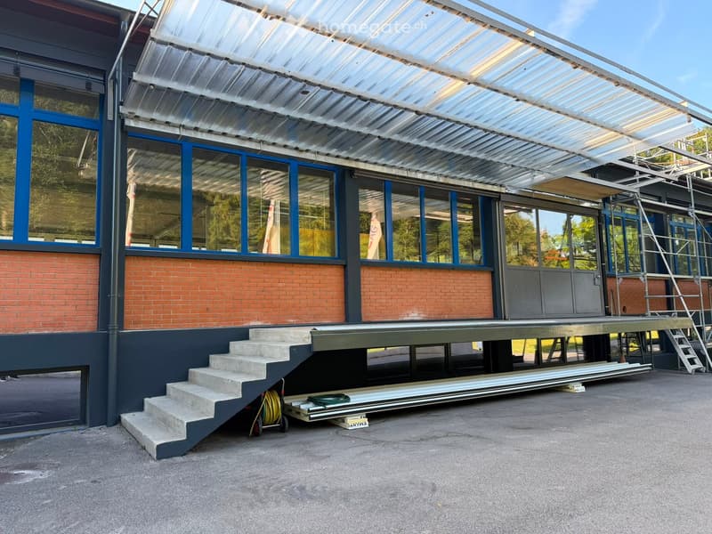 Modern building with a raised ground floor, brick facade, large windows, and a covered walkway leading to a raised platform.