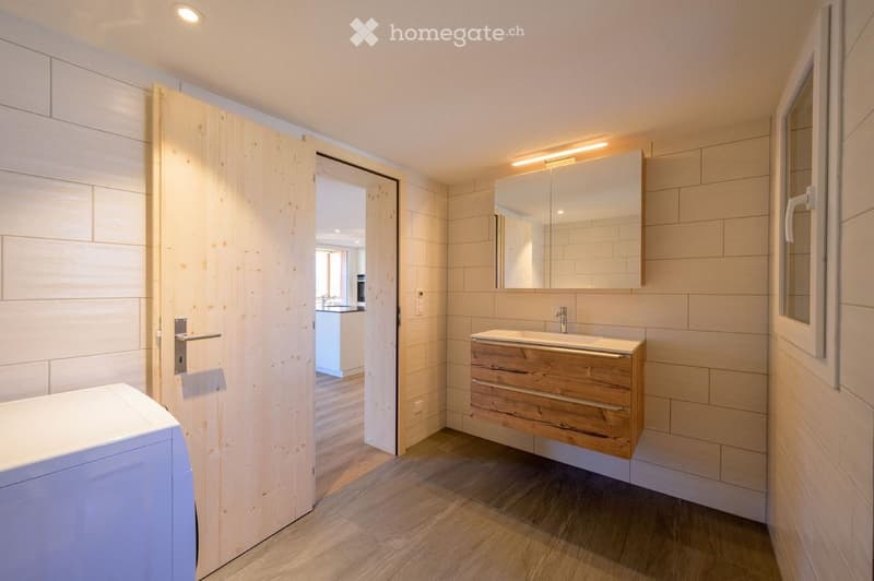 Modern bathroom with light wooden cabinet, sink, and mirror. The room features light-colored tiles and a wooden door leading to another space.