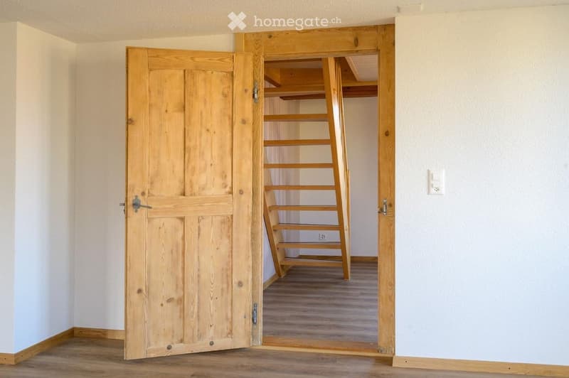 An empty room with wooden flooring and white walls, featuring an open wooden door leading to a closet with wooden shelves and a small ladder.