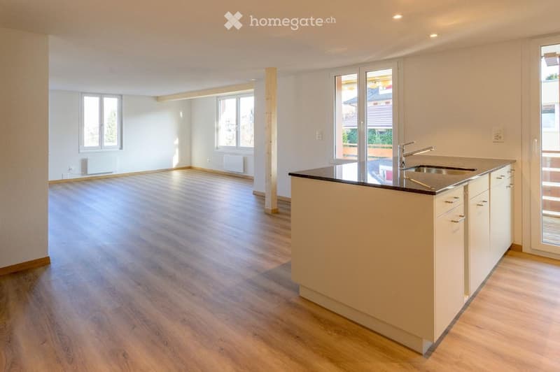 Empty kitchen with light wooden flooring, white walls, and a modern kitchen island with a sink and faucet. Large windows provide natural light.