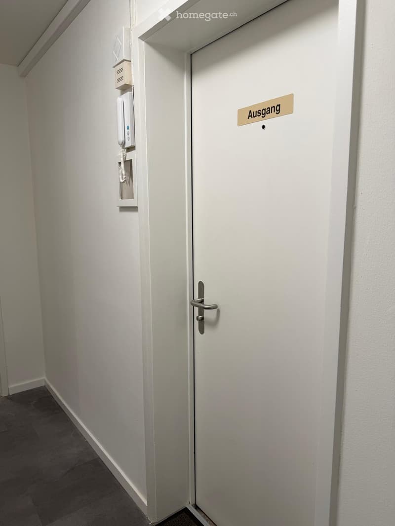A narrow hallway with white walls, a closed door labeled "Ausgang" (exit), and a wall-mounted intercom system.