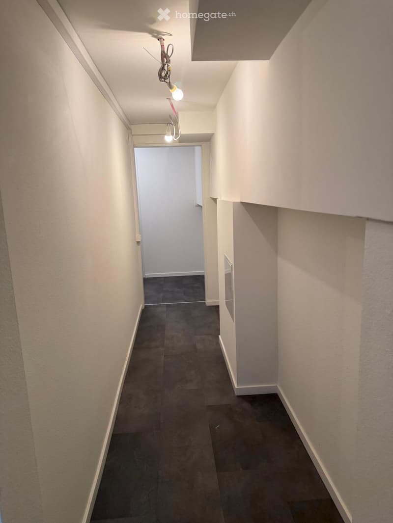Empty hallway with white walls, dark tiled floor, and exposed electrical wiring with light fixtures.