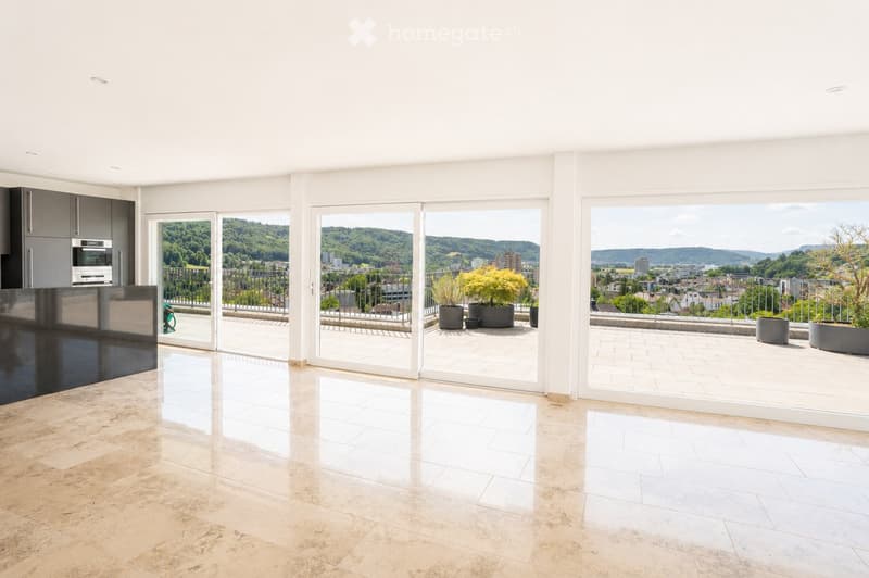 Modern, spacious room with large sliding glass doors leading to a balcony with a scenic city view. The room features polished tile flooring, sleek kitchen cabinets, and an oven visible in the background.