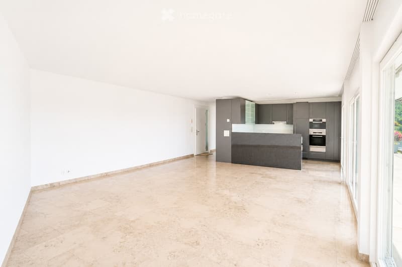 Modern, open-plan kitchen and living area with light-colored tiles, white walls, and grey cabinetry.
