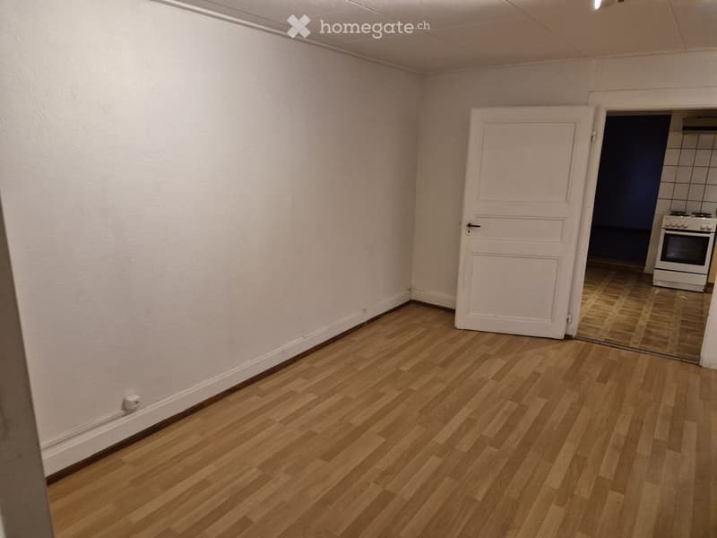 Empty room with light wooden flooring and white walls, featuring a white door leading to another room with a visible oven.