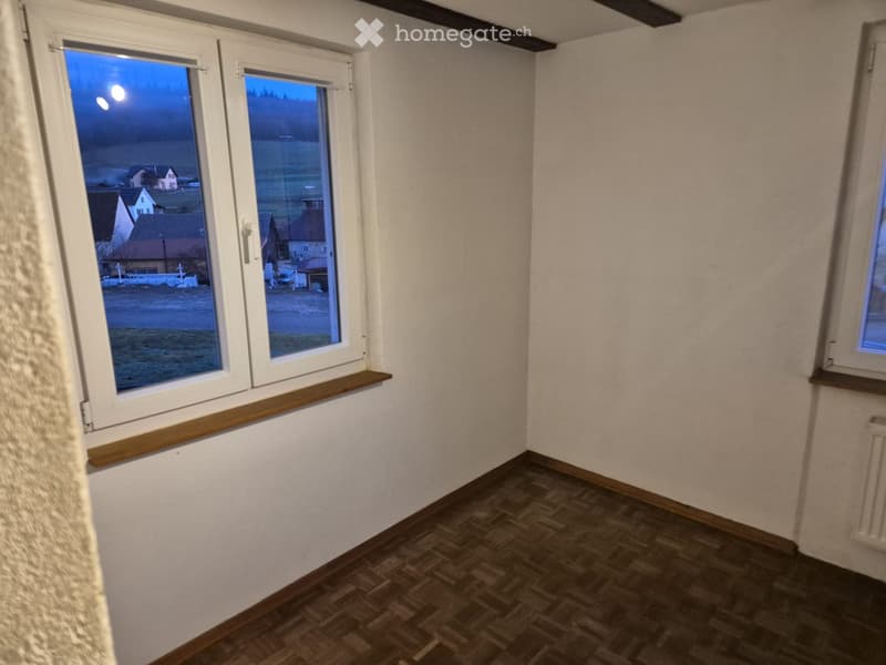 Empty room with wooden flooring, white walls, and large windows showing a view of the countryside at dusk.