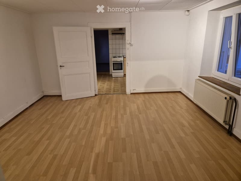 Empty room with wooden flooring, white walls, a white door leading to a kitchen area, and a window with blue glass panes.