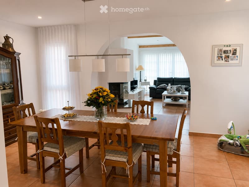 Dining area with a wooden dining table and chairs, a sofa in the living area, a fireplace, a lamp, and a decorative cabinet.