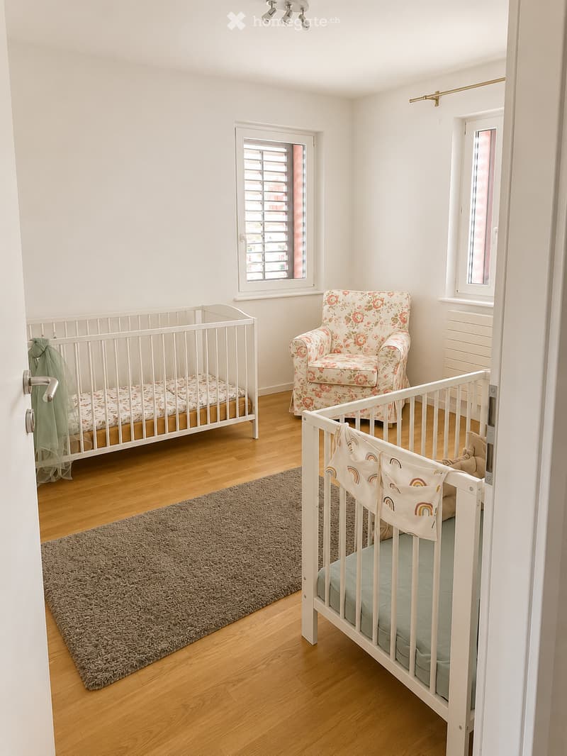Brightly lit nursery with wooden flooring, two cribs, a floral-patterned armchair, and a soft grey rug.