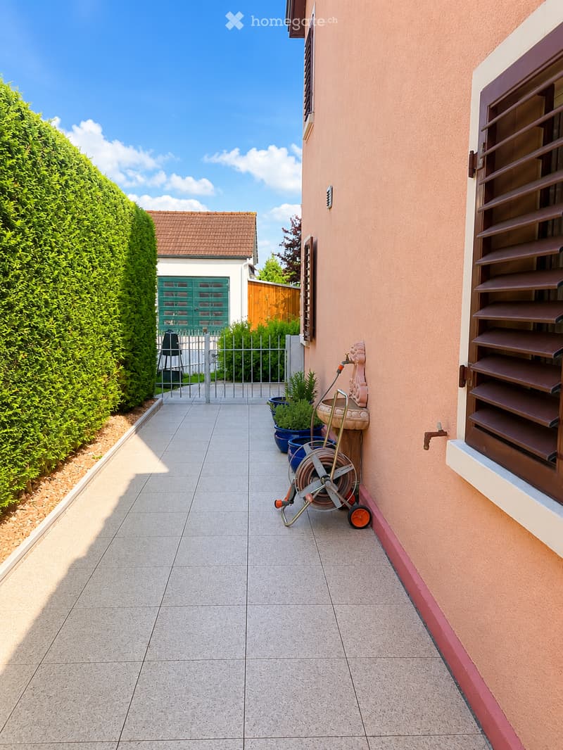 A narrow outdoor pathway leads to a gated entrance, flanked by a neatly trimmed hedge and a building with pink walls and brown shutters. A coiled hose and potted plants are visible along the pathway.
