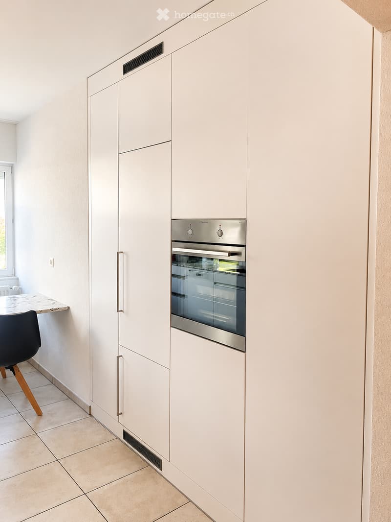 Modern kitchen with white cabinets, stainless steel oven, marble countertop, and a black chair with wooden legs.