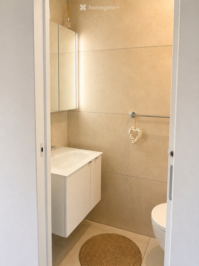 Modern bathroom with beige tiles, white sink cabinet, toilet, and a wicker basket on the floor.