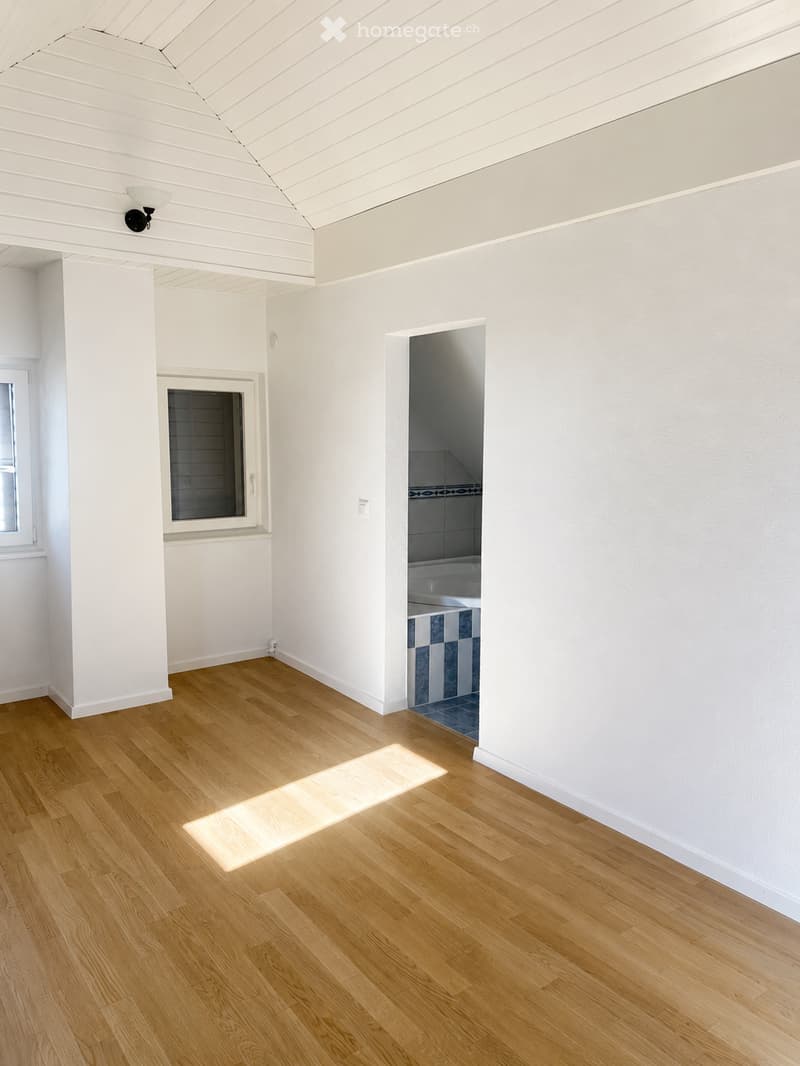 Bright, empty room with light wooden flooring, white walls, and a visible doorway leading to a tiled bathroom area.