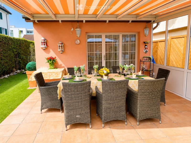 A well-furnished patio with a dining table, six wicker chairs, and decorative vases. The patio features a tiled floor, a covered awning, and a view of a neatly trimmed garden.