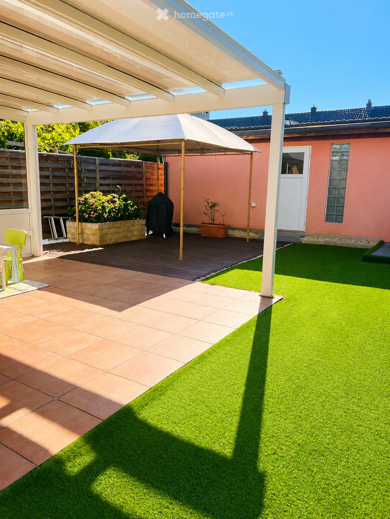 Well-maintained garden with artificial grass, wooden deck, patio umbrella, potted plants, and a covered seating area with a table and chairs.