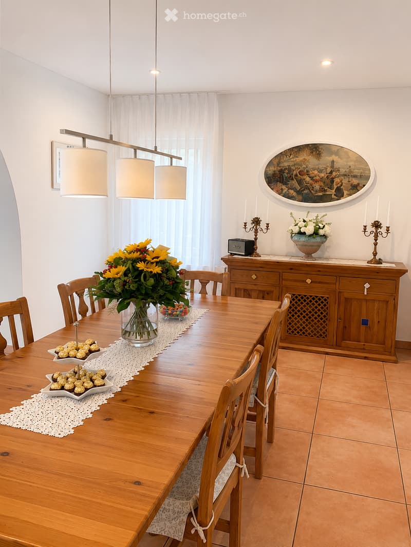 Dining room with a wooden dining table, chairs, a flower vase, and a cabinet with decorative items.
