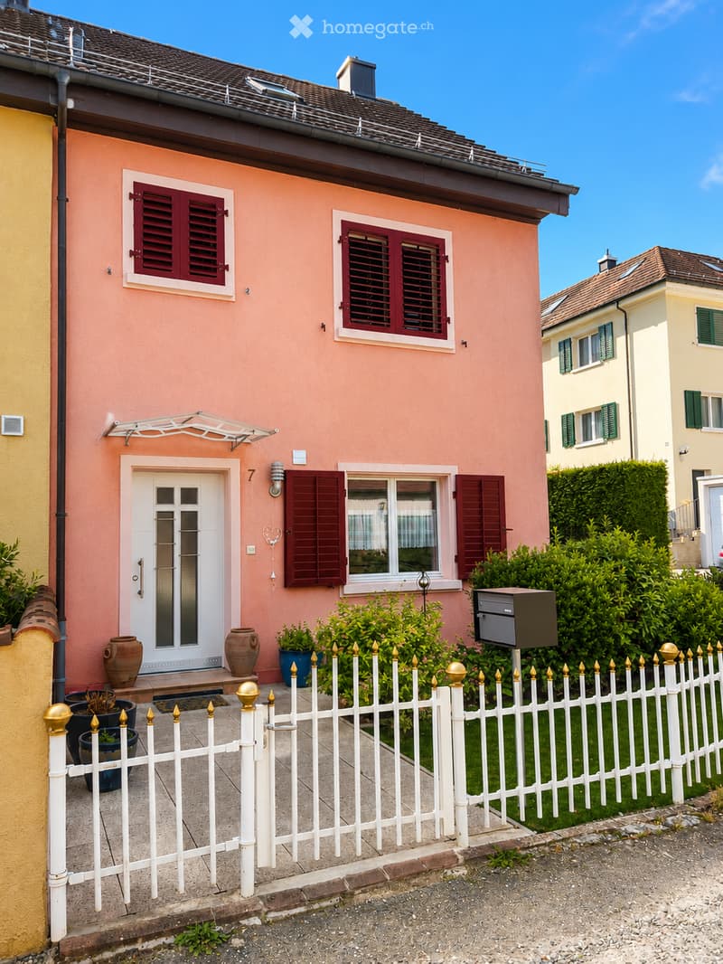 Pink single-story house with white door, red shutters, and decorative vases near the entrance. White picket fence with gold finials surrounds the property.